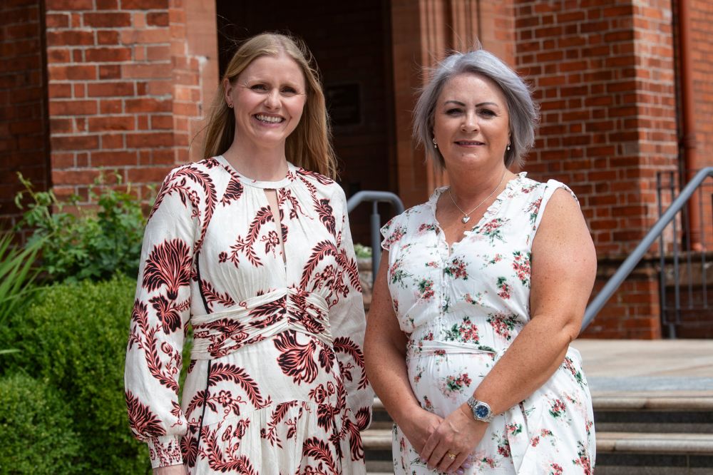 Three female conference attendees in white floral summer dresses posing for a picture in front of a doorway into a red brick building
